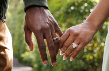 husband and wife holding hands in front of a plant, highlighting the her engagement ring and wedding band and his wedding band