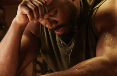 A close-up image of a man with a muscular build, wearing a sleeveless shirt and a cross necklace. He appears to be in deep thought or distress, with his head bowed and one hand resting on his forehead. The lighting is warm and casts shadows, emphasizing the sweat on his skin. The background is blurred and dimly lit, creating an intense and contemplative mood. Beveled Cross Pendant in Sterling Silver This sterling silver cross pendant makes a great gift or daily accessory. It features a stylish beveled edge and comes complete on a matching box chain with a secure lobster clasp.