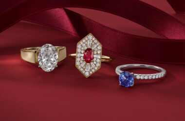 The image showcases three exquisite rings against a rich, red background, with a luxurious red satin ribbon artfully draped in the background, adding to the sense of elegance and sophistication. The rings are as follows: On the left, there is a gold ring with a prominent, oval-shaped clear gemstone, likely a diamond. The gemstone is set in a simple and classic gold band, emphasizing its size and brilliance. In the center, there is a gold ring featuring an intricate design. It has an elongated hexagonal shape with a central red gemstone, possibly a ruby, surrounded by numerous smaller clear gemstones arranged in a detailed pattern, creating a striking and ornate piece. On the right, there is a silver or white gold ring with a round blue gemstone, possibly a sapphire. The gemstone is set in a solitaire style with a band encrusted with small clear gemstones, adding a touch of sparkle and enhancing the overall elegance of the ring. The overall presentation of the rings, combined with the rich red background and ribbon, suggests they are luxurious and high-quality pieces, likely intended for special occasions or as statement jewelry.