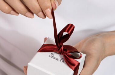 The image shows a close-up of a person untying a red satin ribbon on a small white gift box. The box features the "Shane Co." logo prominently printed on it, indicating that it is from the jewelry company. The person's hands are carefully holding the box and pulling the ribbon to open it. The background is plain and light-colored, which keeps the focus on the hands and the gift box. The overall scene suggests an elegant and special moment, likely involving the unwrapping of a jewelry gift, adding a sense of anticipation and excitement.