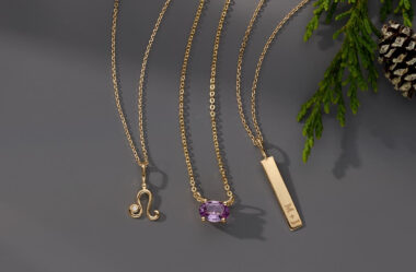 The image features three elegant gold necklaces displayed against a dark, neutral background, with some greenery and a pinecone adding a festive or natural touch to the setting. Each necklace has a unique pendant, showcasing different styles and designs: Left Necklace: This necklace has a delicate gold chain and features a pendant in the shape of a stylized symbol, possibly a zodiac sign (Leo). The pendant includes a small, round clear gemstone, adding a touch of sparkle to the design. Middle Necklace: The middle necklace also has a fine gold chain and displays a prominent oval-shaped gemstone as its pendant. The gemstone is a light purple color, likely an amethyst, and is set in a simple, four-prong setting that allows the stone to be the focal point. Right Necklace: The third necklace has a slightly thicker gold chain and features a rectangular gold bar pendant. The bar pendant is engraved with "M + J," suggesting a personalization option, making it a meaningful piece that could symbolize initials or a special bond between individuals. The overall presentation highlights the variety and versatility of the jewelry pieces, with each necklace offering a distinct look. The background and natural elements enhance the elegance and charm of the necklaces, making them suitable for various occasions and personal styles.