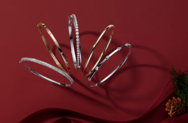 The image showcases a collection of elegant bangle bracelets, artfully arranged against a rich, red background. The bracelets are displayed with a festive touch, including a satin ribbon and a pinecone with some greenery in the lower right corner. The bracelets feature various designs, materials, and styles, highlighting their distinct characteristics: Leftmost Bracelet: This bangle is made of white gold or platinum and is encrusted with small, clear gemstones all around, creating a continuous line of sparkle. The design is luxurious and eye-catching. Second from the Left: This bracelet is crafted from yellow gold and features a design with evenly spaced small diamonds set into the band. The outer edge of the bangle has a decorative, intricate pattern, adding a touch of detail and sophistication. Middle Bracelet: This bangle is a classic white gold or platinum design with a row of clear diamonds set along the top half. The diamonds are round-cut and closely set, creating a dazzling effect. Second from the Right: This bracelet is a simple and elegant yellow gold bangle with a smooth, polished finish. It has a minimalist design, emphasizing the beauty of the gold itself without additional embellishments. Rightmost Bracelet: This bangle is made of white gold or platinum and features a series of bezel-set diamonds evenly spaced around the band. The design is modern and sleek, with the diamonds providing a subtle sparkle. The overall presentation emphasizes the variety and elegance of the bangle bracelets, showcasing their craftsmanship and luxurious appeal. The rich red background and festive elements enhance the visual impact, making the bracelets suitable for special occasions and holiday gifting.